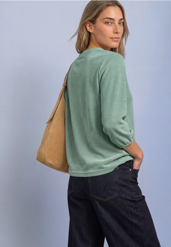 Eine Frau in lässigem grünem Pullover, Jeans und brauner Schultertasche blickt über die Schulter.