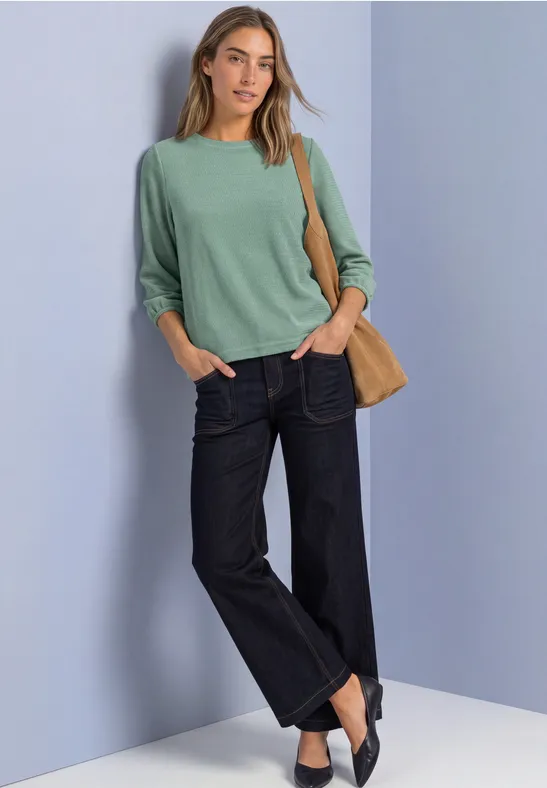 Frau mit mintgrünem Pullover, weiten Jeans, schwarzen Slippern und beigem Shopper vor lilafarbenem Hintergrund.