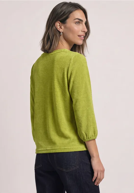 Frau mit schulterlangem dunklem Haar, trägt einen limonengrünen Strickpullover und dunkle Jeans, leicht seitlich fotografiert.