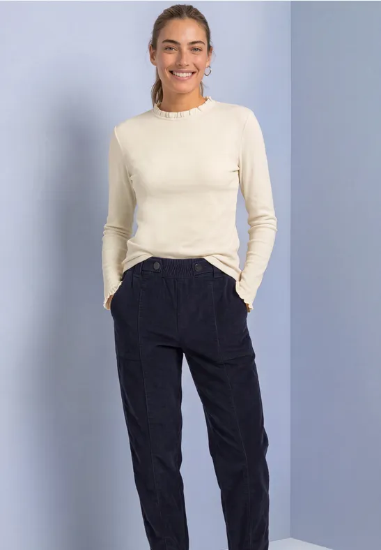 Vrouw lacht, draagt crèmekleurig lang shirt en zwarte broek, voor een lichtblauwe achtergrond.