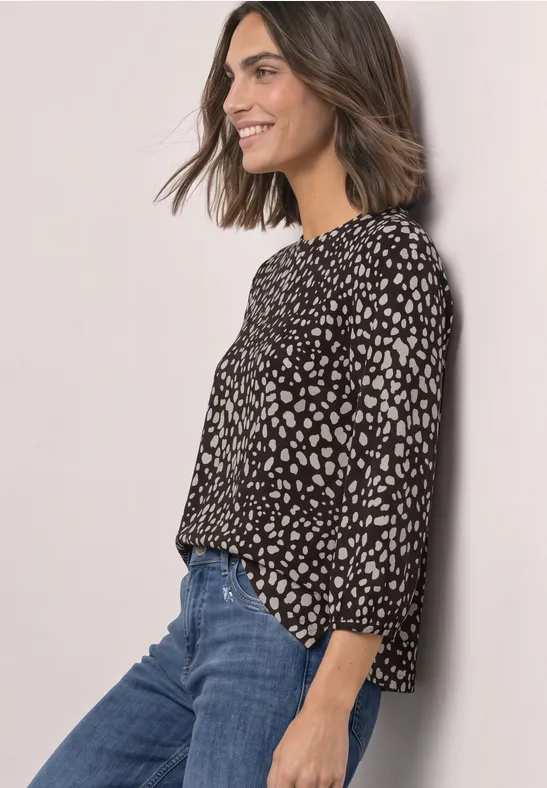 Frau mit Schulterlangem Haar, die seitlich lächelnd eine schwarze Bluse mit weißem Herzmuster und blaue Jeans trägt.