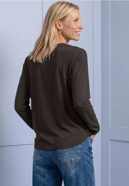 Eine blonde Frau trägt ein schwarzes Langarmshirt und Jeans, steht seitlich vor einer hellblauen Wand.