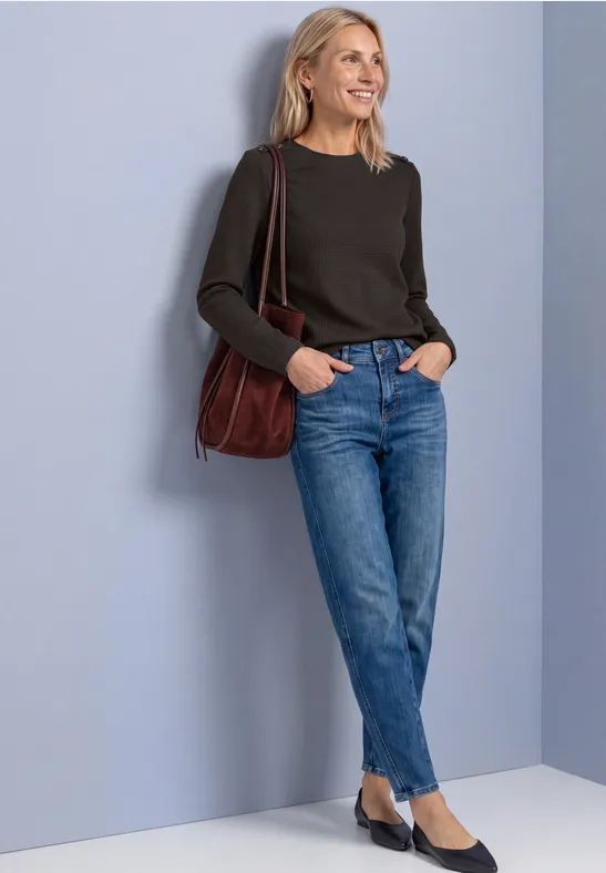 Frau lehnt entspannt an Wand, trägt schwarzen Pullover, blaue Jeans und rote Tasche, lächelt leicht.
