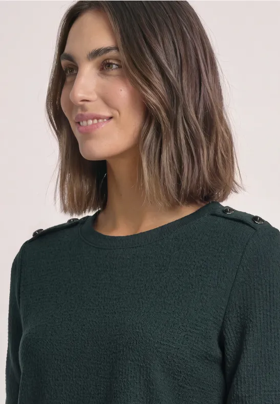 Een glimlachende vrouw met bruin haar draagt een donkergroene, gestructureerde trui met knopen op de schouders.