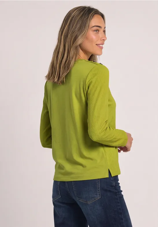 Frau in grünem Langarmshirt und Jeans zeigt sich seitlich, lächelnd.