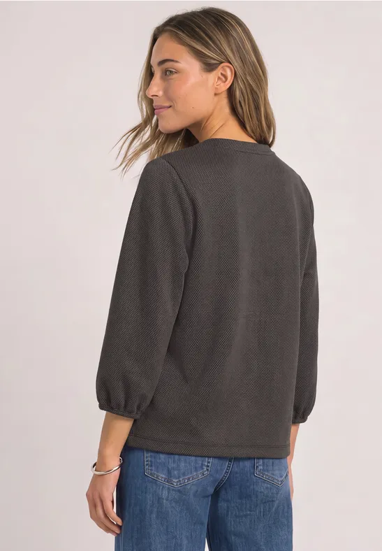 Eine Frau von hinten in dunkelgrauem Pullover mit dreiviertelarm und blauer Jeans.