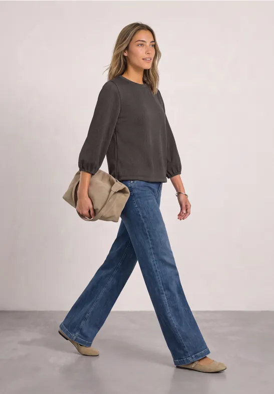 Frau in grauem Pullover, weiten Jeans und beiger Tasche, läuft in schlichtem Studiohintergrund.