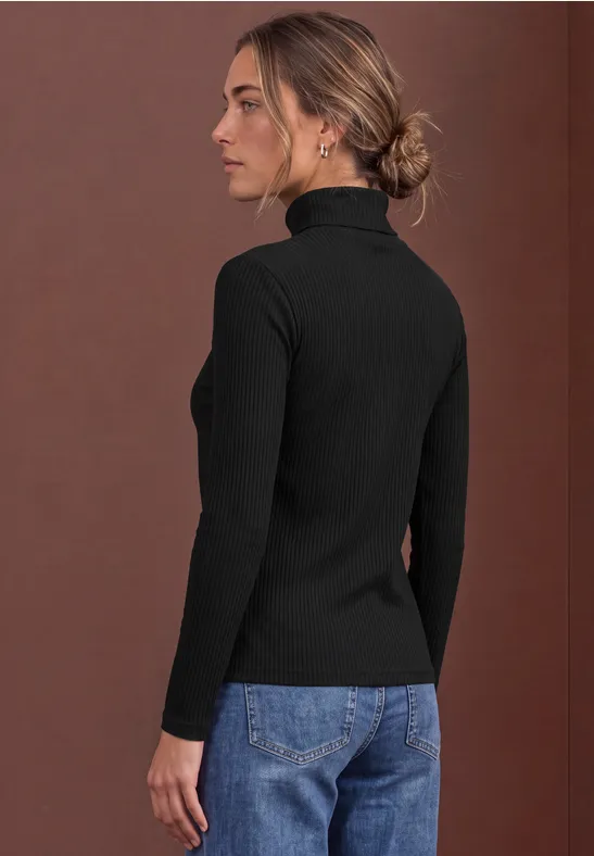 Eine Frau in schwarzem Rollkragenpullover und Jeans steht seitlich vor braunem Hintergrund.