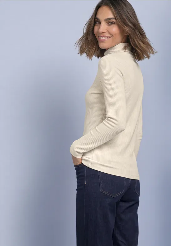 Frau in cremefarbenem Rollkragenpullover und blauen Jeans schaut über die Schulter und lächelt.