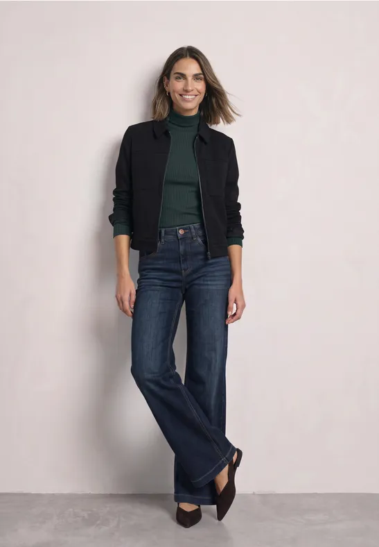 Eine Frau in schwarzer Jacke, grünem Oberteil und weiten Jeans steht vor hellem Hintergrund und lächelt.