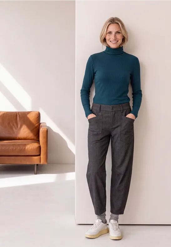 Vrouw met kort blond haar leunt tegen een witte muur, draagt groene coltrui, grijze broek en witte sneakers.