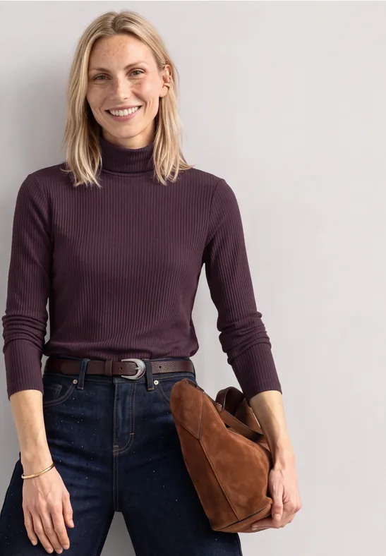 Frau mit blondem Haar, dunkelviolettem Rollkragenpullover und Jeans, hält eine braune Tasche und lächelt.