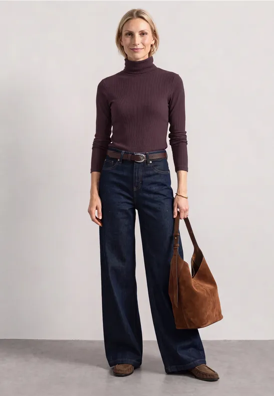 Frau im lila Rollkragenpullover mit weiten Jeans, braunem Gürtel, Tasche und Loafers.