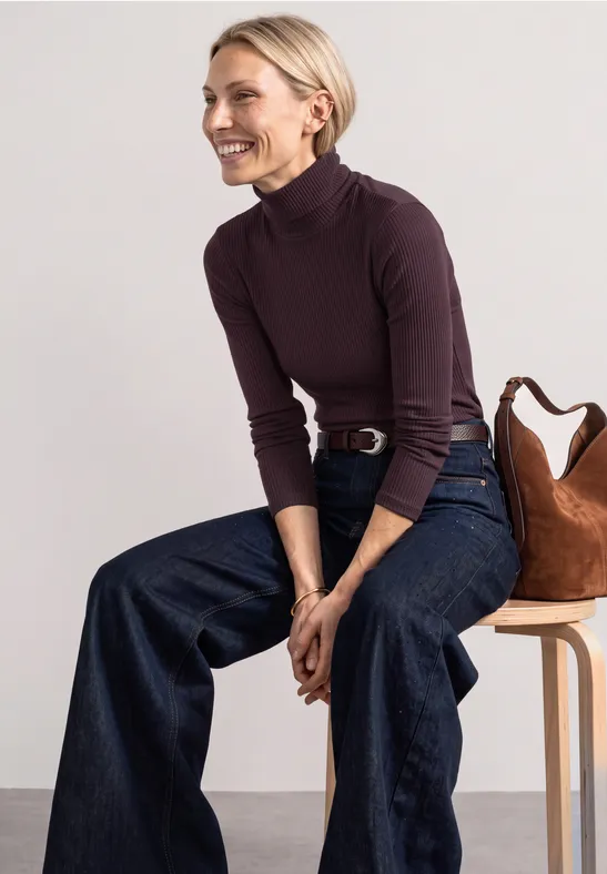 Frau in lila Rollkragenpullover und weiten Jeans sitzt lächelnd auf einem Hocker mit Tasche.