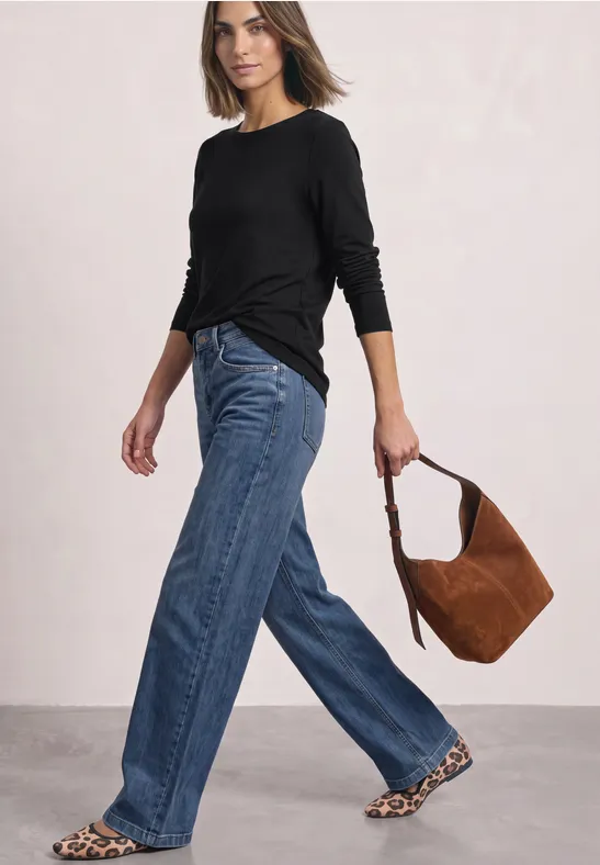 Frau mit schwarzem Longsleeve, weiten Jeans und Leoparden-Ballerinas, hält eine braune Wildleder-Tasche.