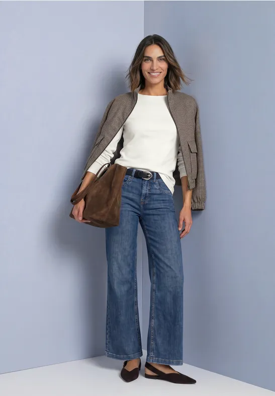 Femme souriante en jean évasé, top blanc, veste taupe et sac marron devant un fond gris.