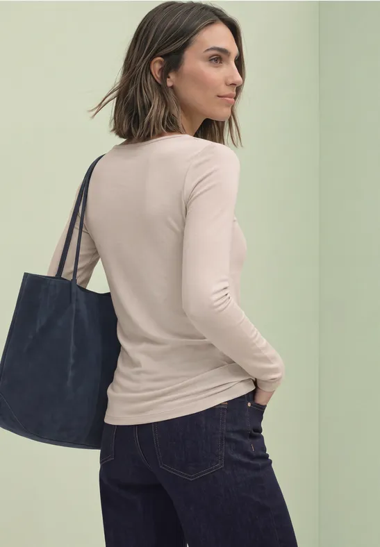 Frauenrückansicht in beiger Bluse, dunklen Jeans und dunkelblauer Tasche vor pastellgrüner Wand.
