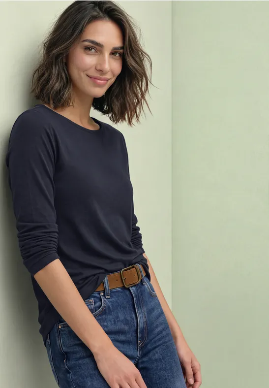 Eine lächelnde Frau in dunkelblauem Longsleeve und Jeans lehnt entspannt an einer hellen Wand.