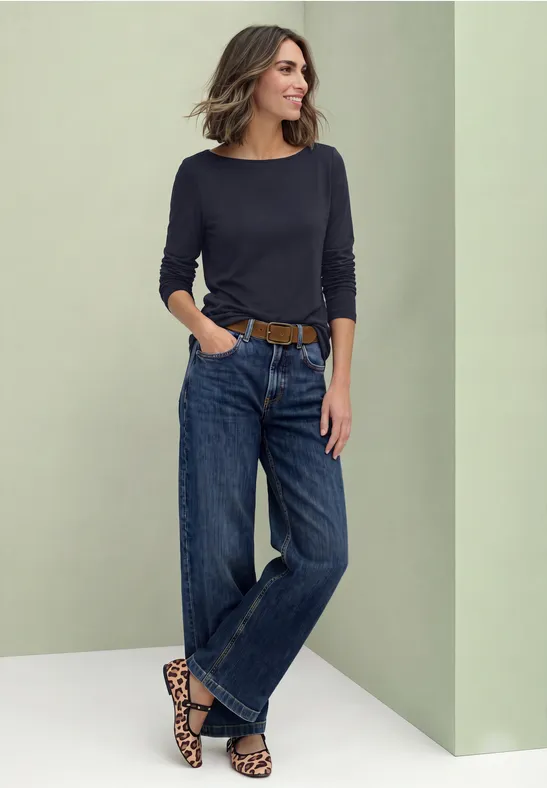 Eine Frau lehnt an einer Wand, trägt schwarzen Pullover und weite Jeans mit Leoparden-Gürtel.