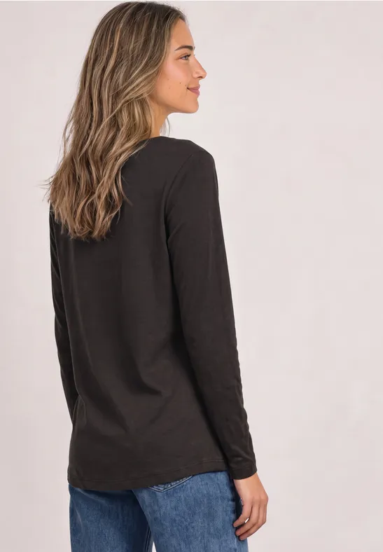 Frau steht seitlich, trägt schwarzes Langarmshirt und Jeans vor hellem Hintergrund.