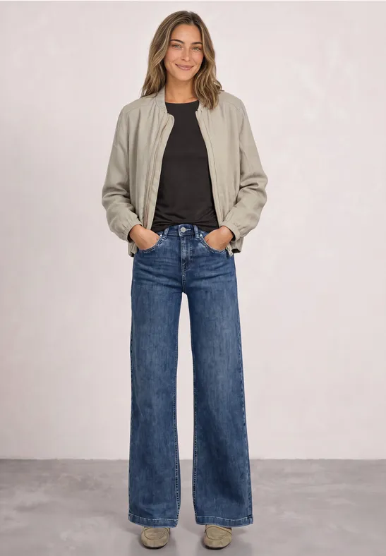 Frau mit beige Bomberjacke, schwarzem Shirt und weiten Blue-Jeans vor schlichter Wand.