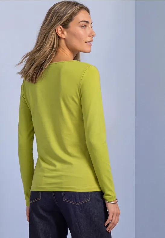 Rückansicht einer Frau in einem hellgrünen Langarmshirt und blauen Jeans vor neutralem Hintergrund.