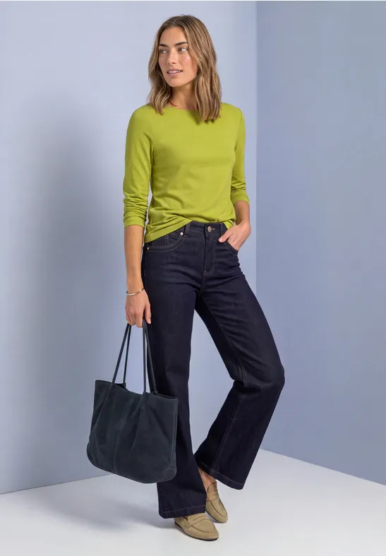 Frau in limonengrünem Langarmshirt, dunklen Jeans und beigen Boots, mit dunkelblauer Tasche, vor hellblauem Hintergrund.