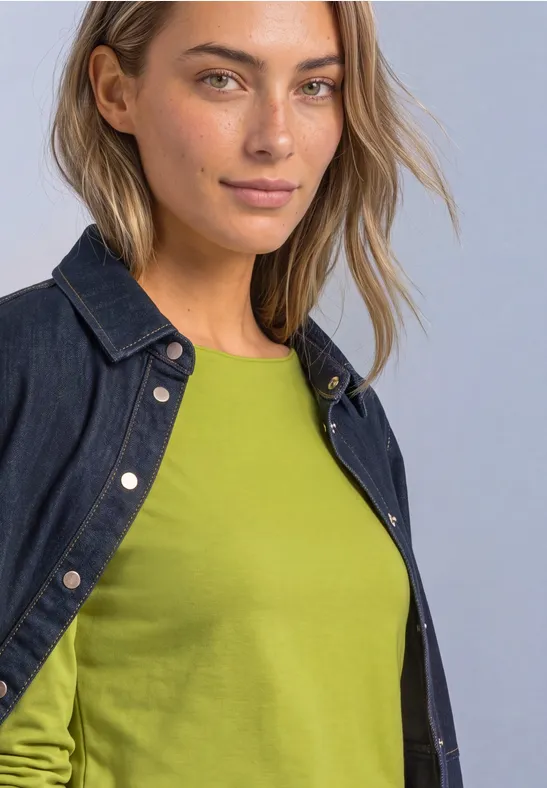 Frau mit kurzem blondem Haar lächelt, trägt dunkelblaue Jeansjacke und grünes Shirt.