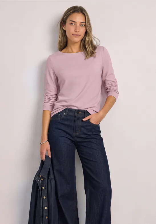 Frau in lockerem rosa Langarmshirt und weitem Jeans, hält eine Jacke in der Hand.
