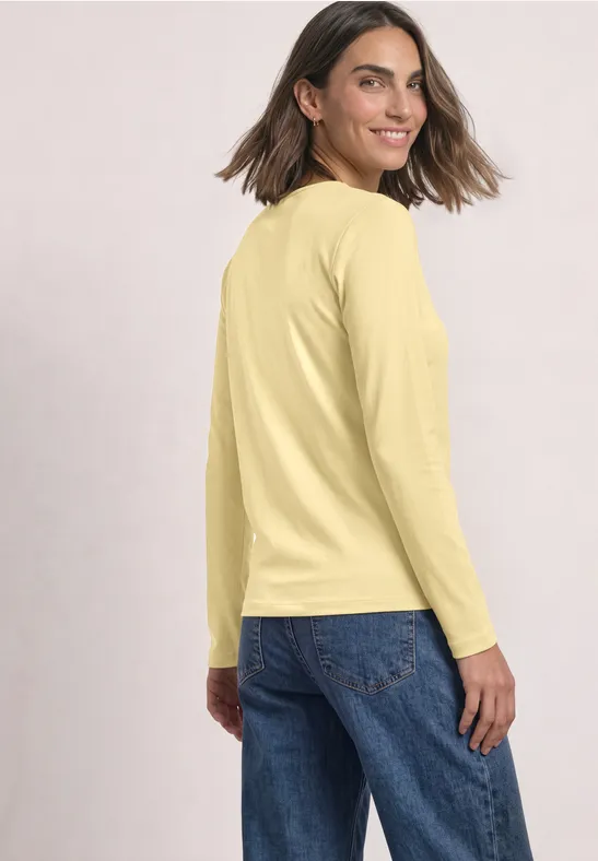 Femme portant un t-shirt jaune à manches longues et un jean bleu, regard par-dessus l’épaule.