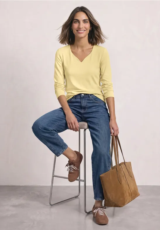 Femme en haut jaune et jean bleu assise sur un tabouret, souriante, tenant un sac marron.