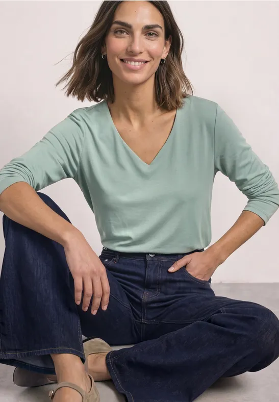 Vrouw zit in kleermakerszit, draagt een groen V-hals top en wijde jeans, lacht ontspannen.