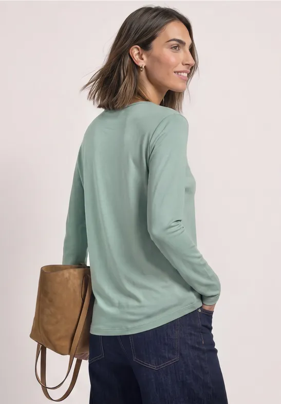 Vrouw met kort bruin haar, gezien van achter, draagt een groen langarmshirt, donkere spijkerbroek en een bruine tas.