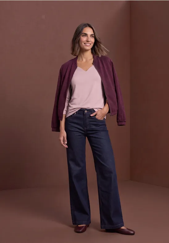 Eine lächelnde Frau in violetter Bomberjacke, rosa Top und weiten, hochgeschnittenen Jeans vor braunem Hintergrund.