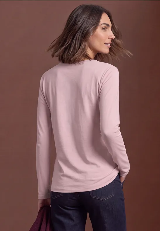 Frau in rosa Langarmshirt und Jeans, leicht von hinten, Blick über die Schulter vor neutralem Hintergrund.