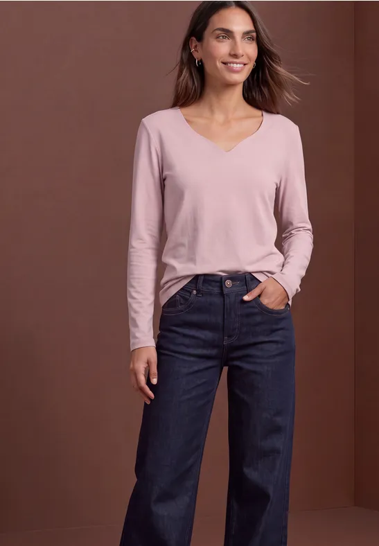 Frau trägt rosa V-Ausschnittshirt und weite dunkle Jeans vor braunem Hintergrund.