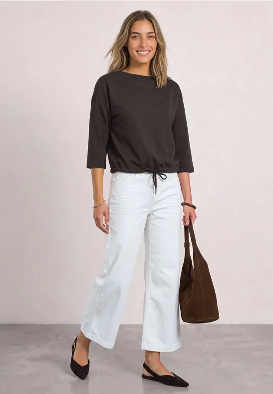 Frau in schwarzem 3/4-Ärmel-Shirt, weißen weiten Jeans und schwarzen Ballerinas, trägt braune Tasche.