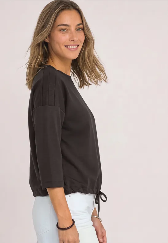 Frau im Profil lacht, trägt schwarzes 3/4-Arm-Shirt mit Kordelzug und weiße Hose vor hellem Hintergrund.