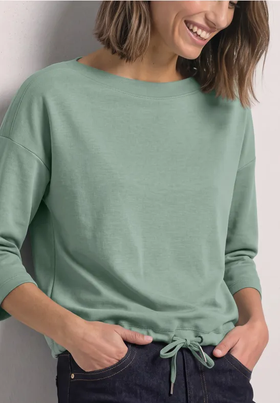 Frau in grünem Dreiviertelarmshirt mit Kordelzug am Saum, Hände in Hosentaschen, lächelt.