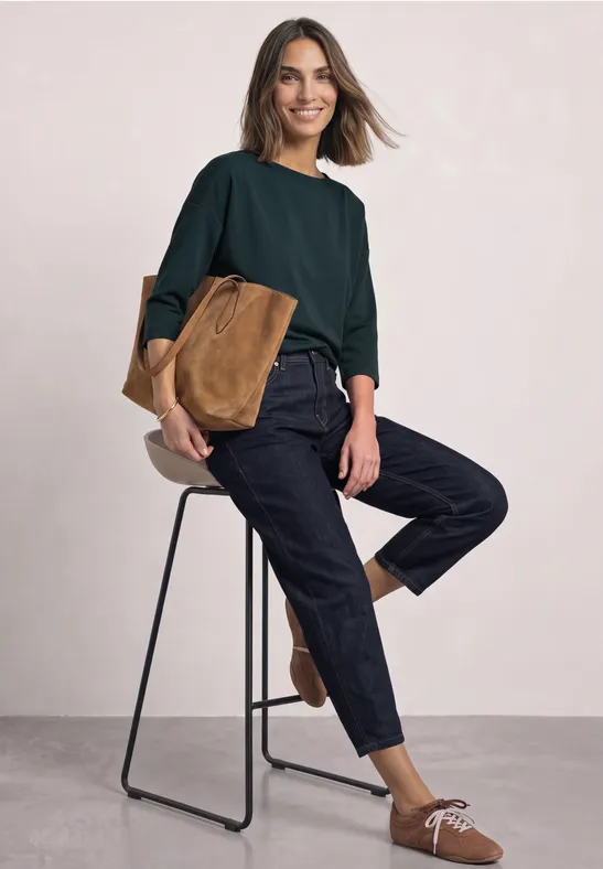Frau in dunkelgrünem Pullover und schwarzer Hose sitzt lässig auf einem Hocker, mit hellbrauner Tasche und Mokassins.
