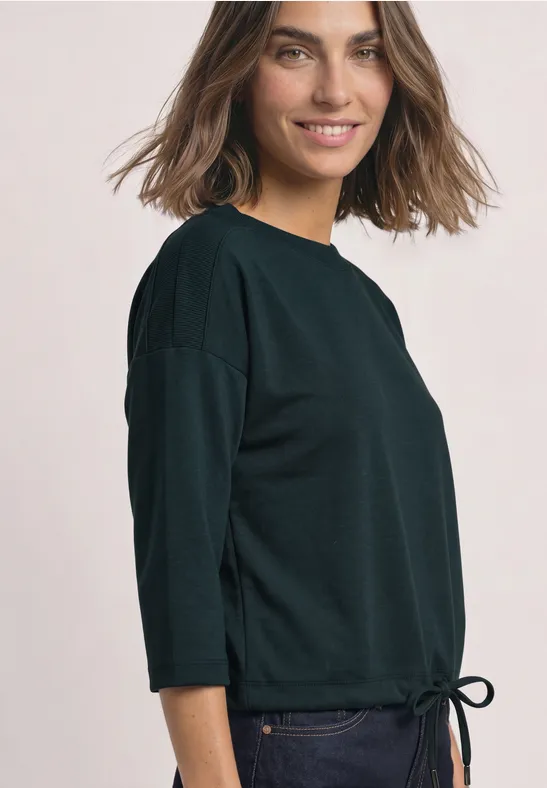 Eine lächelnde Frau im schwarzen 3/4-Arm-Pullover mit Tunnelzug am Saum, in leichter Seitenansicht.