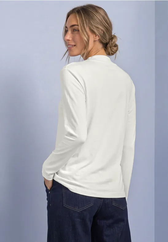 Vrouw gezien van achter, kijkt over schouder, draagt wit shirt met lange mouwen en een donkerblauwe jeans