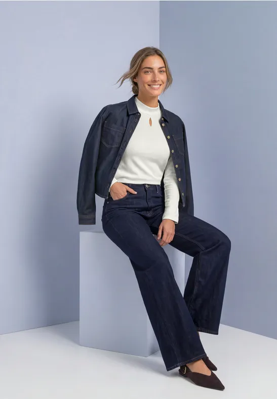 Vrouw in donker denim pak en wit topje, glimlachend zittend op een lichte kubus voor een neutrale achtergrond.
