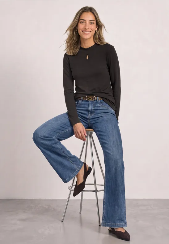 Femme blonde en pull noir et large jean, assise avec décontraction sur un tabouret