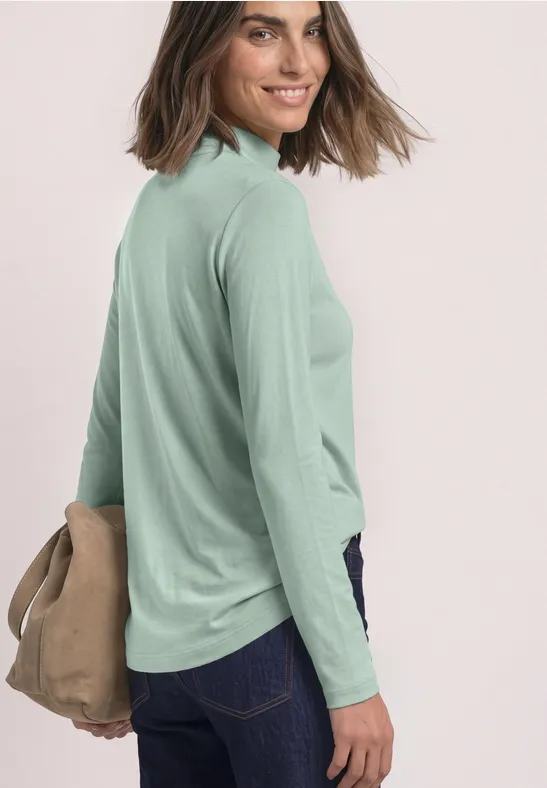 Frau im Profil trägt hellgrünes Langarmshirt, dunkle Jeans, hält eine beige Tasche und lächelt.