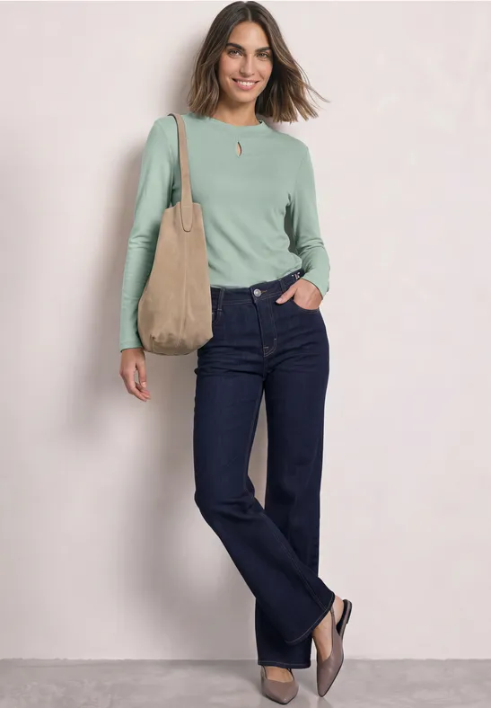 Frau in grünem Langarmshirt, dunklen Jeans, brauner Tasche auf der Schulter, lächelt vor hellem Hintergrund.