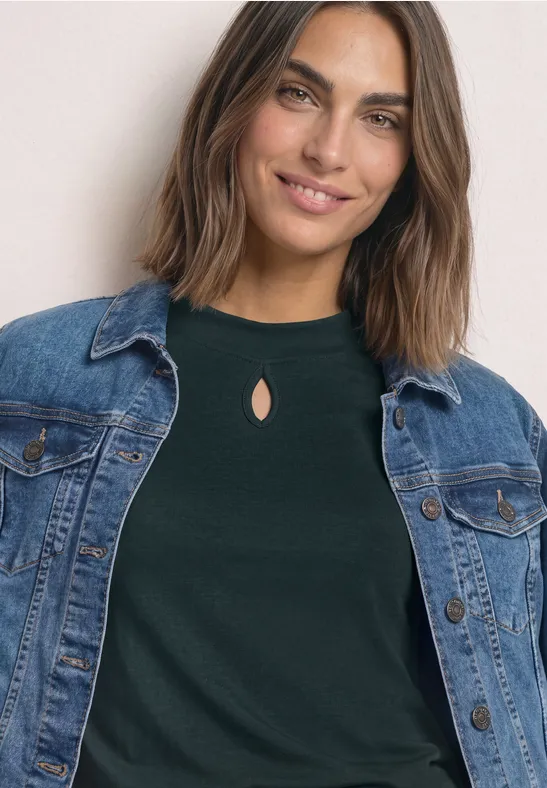 Femme souriante en veste en jean et top vert foncé à encolure fendue, gros plan.