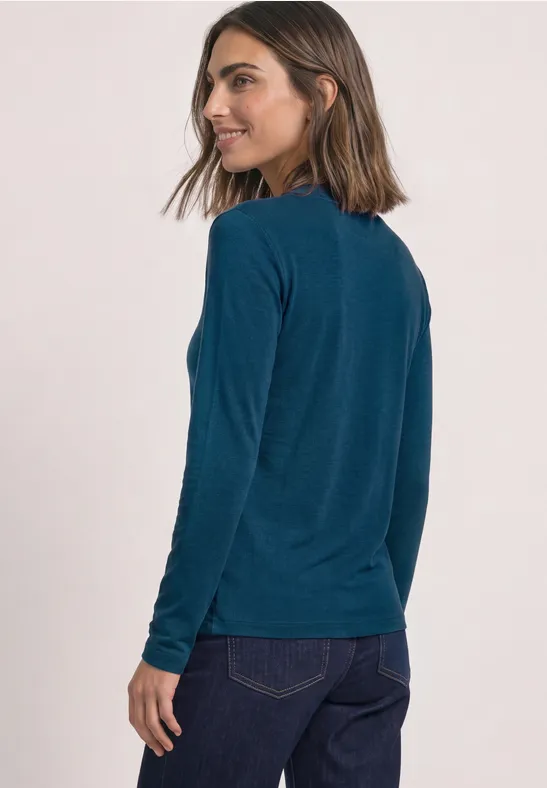 Frau in langärmeligem, petrolfarbenem Shirt und dunkler Jeans, steht seitlich gewandt und lächelt.