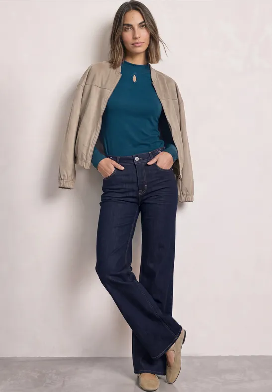 Eine Frau posiert in einer blauen Bluse, dunkelblauen Jeans und einer beigefarbenen Jacke über den Schultern.
