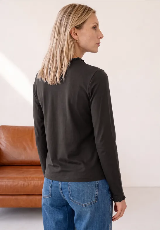 Eine Frau mit blonden Haaren in schwarzem Langarmshirt und blauen Jeans steht seitlich vor einem Sofa.
